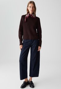 Femme portant un pull côtelé marron foncé, un jean bleu marine à jambes larges, des mocassins noirs et un foulard à rayures bordeaux autour du cou, se tenant devant un fond uni.