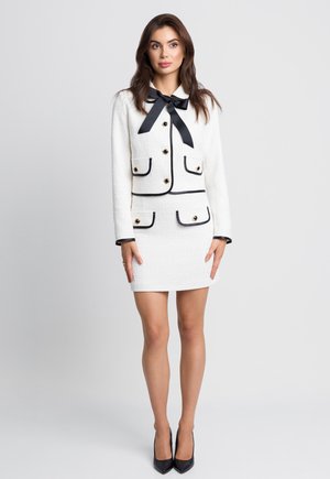 Biały kostium składający się z dopasowanej marynarki z czarnymi wykończeniami i kieszeniami, zestawiony z dopasowaną mini spódnicą. Akcent w postaci kokardy przy kołnierzu.