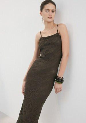 Mlada ženska se naslanja na steno, oblečena v oprijeto temno rjavo čipkasto obleko s tankimi naramnicami in z debelimi zapestnicami na levi roki.