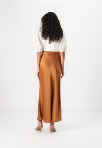 Gonna in satin marrone con uno spacco, abbinata a una blusa bianca testurizzata. L'outfit presenta maniche a palloncino e tacchi nudi minimalisti.