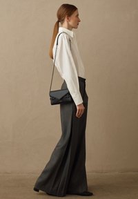 Sac en cuir noir en bandoulière avec un rabat géométrique, porté sur une chemise blanche à boutons et un pantalon taille haute gris. Texture lisse, design minimal.
