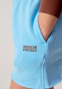 Main dans la poche d'un short bleu clair avec "American Vintage" imprimé en marron sur le tissu, porté avec un haut à manches longues marron.
