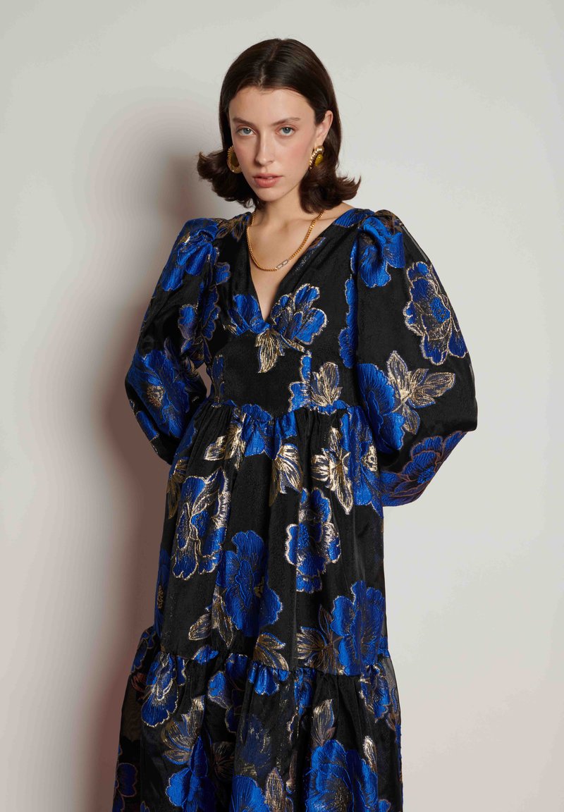 Μαύρο φόρεμα με μπλε και χρυσό λουλουδάτο σχέδιο, μανίκια φουσκωτά, λαιμόκοψη V και στρωματοποιημένη σχεδίαση. Υφασμάτινη υφή που προσθέτει βάθος.
