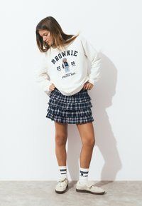 Sweatshirt col rond en blanc cassé avec le texte "BROWNIE" et un graphique d'ours, associé à une jupe mini plissée à carreaux navy ; chaussettes blanches et baskets beiges.