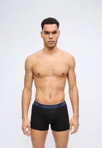 Άνδρας χωρίς πουκάμισο με κοντά, σγουρά μαλλιά, που φοράει μαύρα μποξεράκια Tommy Hilfiger, στέκεται μπροστά σε απλό ανοιχτό γκρι φόντο.