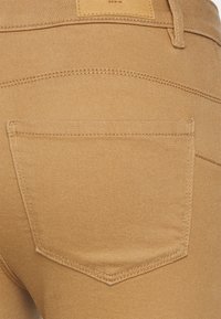 Jeans de denim color tostado con un bolsillo trasero que presenta costuras sutiles. La tela tiene una textura suave y un diseño clásico de cinco bolsillos.
