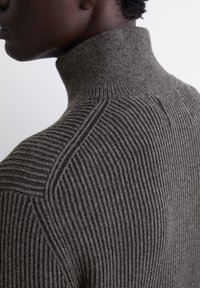 Grauer gerippter Rollkragenpullover mit einer figurbetonten Passform, der einen hohen Kragen und ein strukturiertes Muster aufweist, seitlich betrachtet.