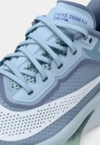 Svetle modré tenisky Nike Zoom Fly vyrobené z kombinace síťoviny a pevných materiálů, s texturovanými akcenty, výrazným bílým swooshem a plochými tkaničkami.