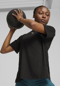 Zwart sportshirt met korte mouwen en een wit logo, gedragen terwijl er een gestructureerde zwarte medicijnbal wordt opgetild tegen een neutrale achtergrond.