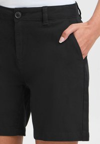 Svarte shorts laget av glatt stoff med knappestengning og sidelommer. Har en skreddersydd passform og ren kant.