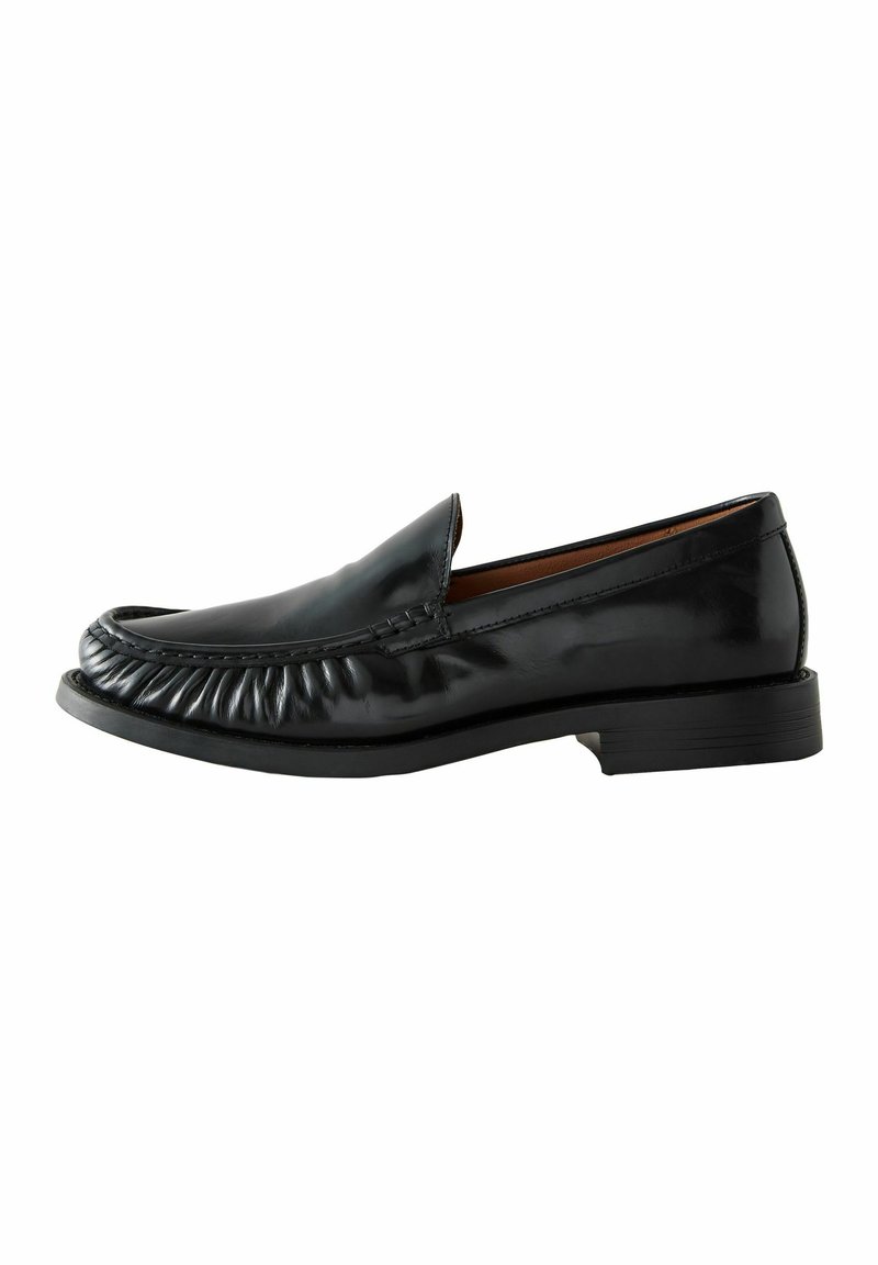 Mocasines de cuero negro con detalles de costura alrededor de la parte superior, punta redondeada y un tacón bajo y texturizado.