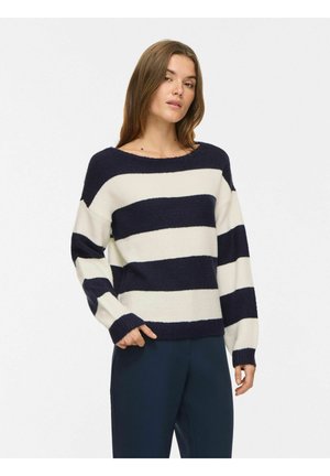 Femme portant un pull à larges rayures horizontales bleu marine et blanc avec des manches longues et un pantalon bleu marine, debout devant un fond clair uni.
