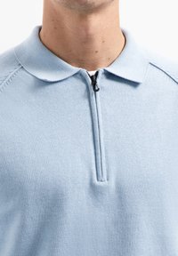 Lichtblauwe poloshirt met een ritssluiting en een zachte, textuurstof. Heeft een klassieke kraag en gebreide details op de schouders.