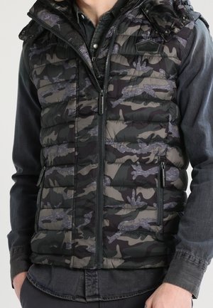 Homme portant un gilet matelassé camouflage noir et gris par-dessus une chemise boutonnée gris foncé, les mains reposant près des poches.