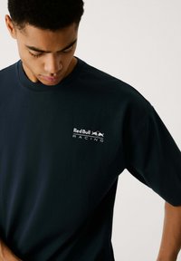 Schwarzes T-Shirt mit kurzen Ärmeln aus Baumwolle, mit einem weißen "Red Bull Racing"-Logo auf der linken Brust. Schlichte, lockere Passform.