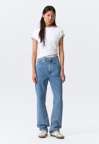 Νεαρή γυναίκα όρθια, φορώντας λευκό t-shirt με γυρισμένα μανίκια, άνετο μπλε τζιν και μπεζ αθλητικά παπούτσια, μπροστά σε απλό ανοιχτό φόντο.