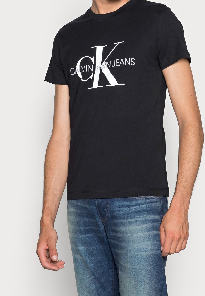 Svart bomull t-shirt med en stor vit Calvin Klein Jeans-logotyp. Korta ärmar, rund hals och en åtsittande design. Bärs med blå denimjeans.