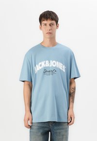Ung mand med kort, mørkt hår iført en lyseblå oversized T-shirt med hvid "JACK&JONES Studio Co." tekst og blå jeans, tatovering på venstre underarm.
