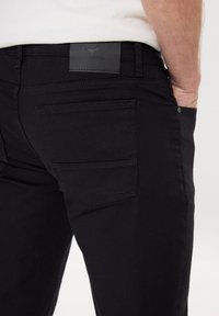 Černé džíny z denimu s texturovanou černou koženou štítkem. Mají jednu zadní kapsu a viditelné švy podél švů.