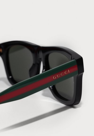 Óculos de sol pretos com hastes listradas em verde e vermelho, apresentando o logótipo dourado "GUCCI". Acabamento liso e brilhante; lentes escuras. Formato retangular.