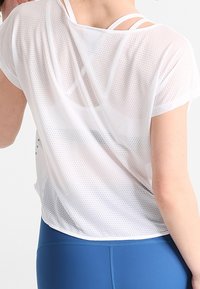 Biała, przewiewna sportowa bluzka z krótkimi rękawami i luźnym krojem, z wycięciem na plecach i podwójnymi, warstwowymi cienkimi ramiączkami pod spodem.