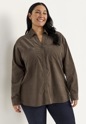 Glimlachende vrouw met lang donker haar draagt een losse bruine blouse met knopen en donkere spijkerbroek tegen een effen lichte achtergrond.