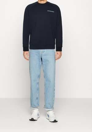 Mörkblå sweatshirt med ribbade muddar och fåll, med vit text "SCOTCH & SODA" på bröstet, kombinerad med ljusblå jeans och vita sneakers.