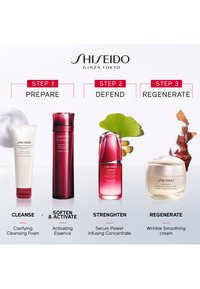 Vier huidverzorgingsproducten horizontaal gerangschikt: een witte tube, een hoge rode fles, een korte rode fles en een wit potje met een gouden deksel.