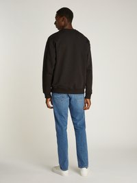 Sudadera negra, de cuello redondo, mezcla de algodón, corte holgado. Combinada con jeans azul claro y zapatillas blancas, de pie contra un fondo neutro.
