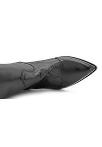 Bota de tobillo de cuero negro con puntera afilada, con costuras decorativas y acentos recortados a lo largo del caña. Textura suave, diseño elegante.