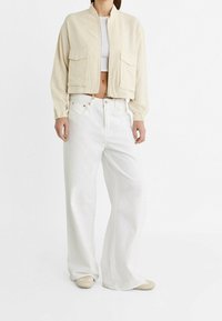 Chaqueta corta beige con dos bolsillos frontales, puños elásticos y un cuello alto, combinada con vaqueros blancos de corte holgado y zapatos beige claro.