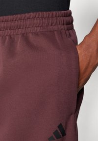 Bordeaux sportbroek met een zachte textuur, elastische tailleband, zijzak en een zwart logo-accent aan de onderkant aan de linkerkant.