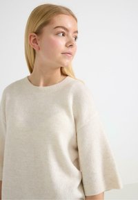 Beżowy, dziewiarski sweter z szerokim okrągłym dekoltem i krótkimi rękawami, charakteryzujący się miękką fakturą i subtelnymi różnicami w odcieniu kolorów.