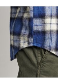 Camicia di flanella a scacchi blu, bianca e nera, con orlo curvo, abbinata a pantaloni verde oliva con cuciture e texture visibili.