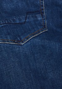Primer plano de una tela de denim azul que muestra la esquina de un bolsillo trasero cosido con detalles en hilo naranja.