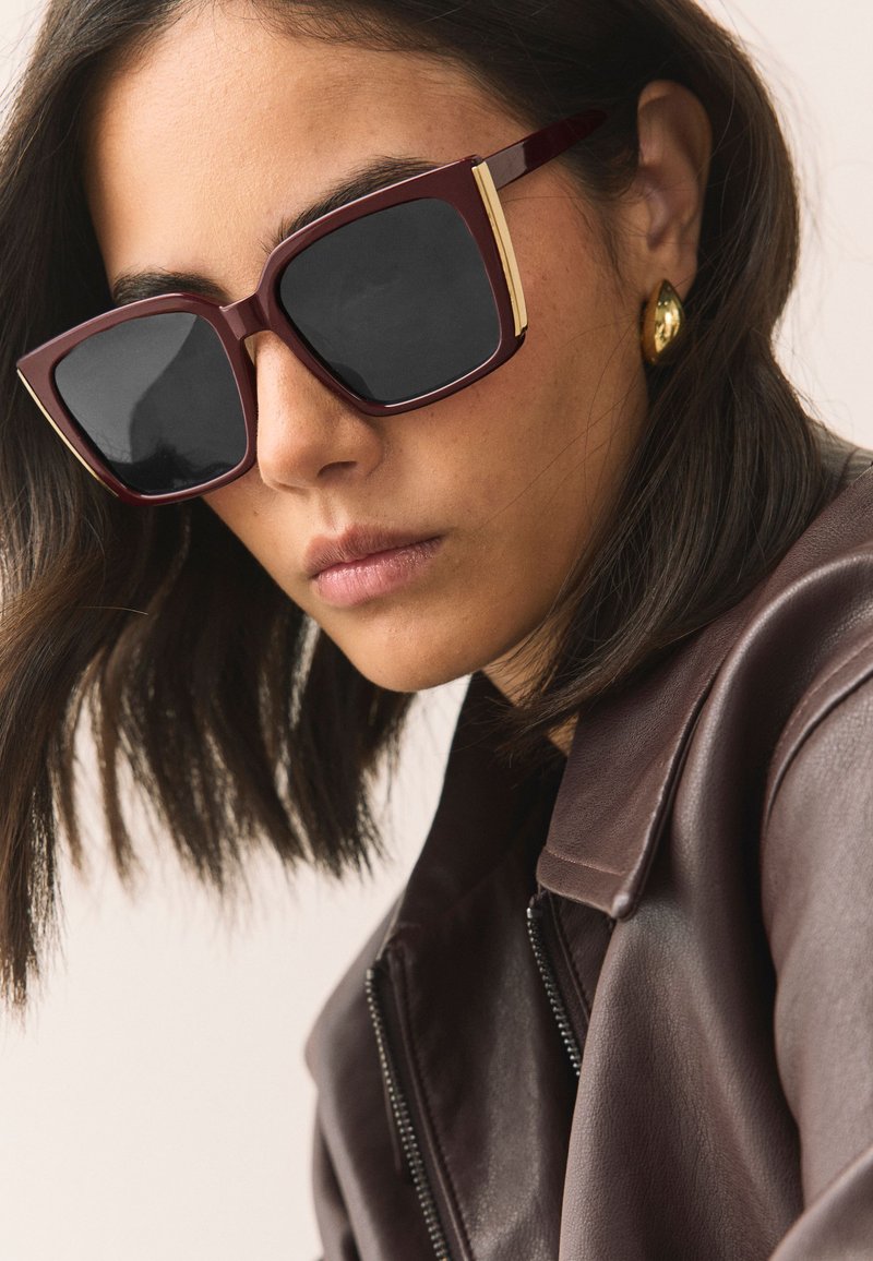 Nainen, jolla on tumma tukka, suuria neliönmuotoisia viininpunaisia aurinkolaseja, kultaiset korvakorut ja ruskea nahkatakki, katsoo hieman alas.