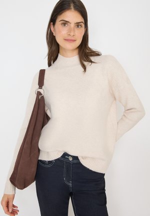 Helles beige Strickpullover mit geripptem Kragen und Bündchen, kombiniert mit dunkelblauen Jeans. Eine braune Wildleder-Schultertasche mit einem Ringakzent ist sichtbar.
