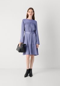 Abito lungo a maniche lunghe di lavanda con corpetto arricciato, vita stretta e gonna A-line fluida. Abbinato a una borsa nera e stivaletti alla caviglia.