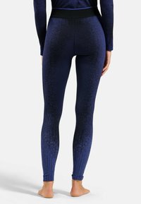 Frau in nahtlosen, navyblauen und schwarzen strukturierten Leggings und passendem Oberteil, barfuß stehend mit dem Rücken zur Kamera.