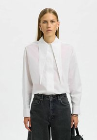 Naine, kellel on sirged blondid juuksed, kannab valget pluusi pika terava kraega ja tumedaid kõrge vöökohtadega teksaseid, hoides musta käekotti.