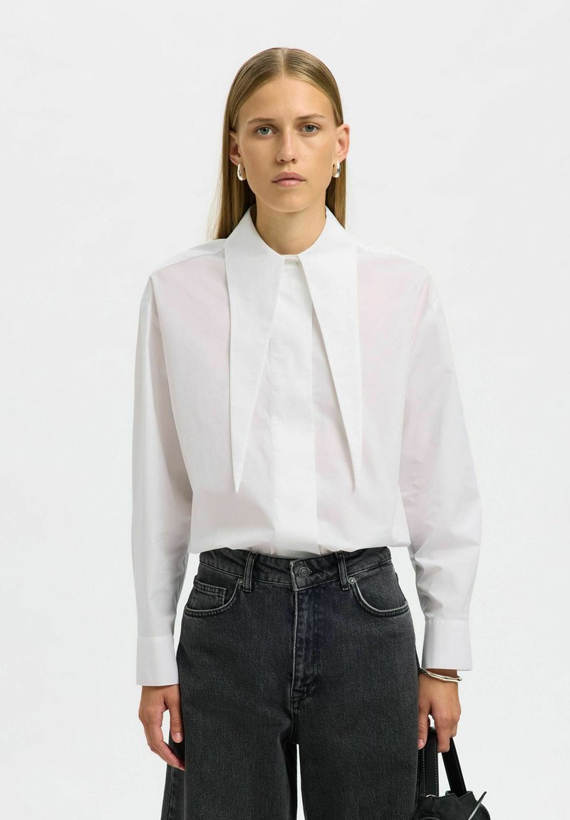 Naine, kellel on sirged blondid juuksed, kannab valget pluusi pika terava kraega ja tumedaid kõrge vöökohtadega teksaseid, hoides musta käekotti.