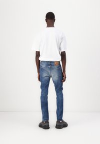 Man som står vänd mot en vit vägg, klädd i en vit t-shirt, blå jeans med en synlig brun läderlapp och svarta sneakers.