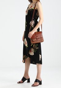 Kobieta ubrana w czarną sukienkę w kwiaty zapinaną na kokardę, czarne sandały na obcasie z odkrytymi palcami oraz nosząca brązową skórzaną torebkę na ramię.