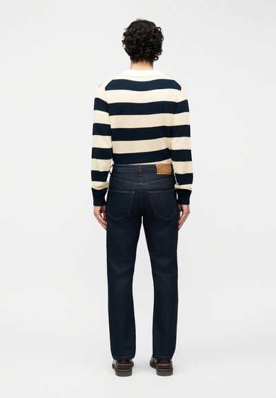 Άτομο με κατσαρά μαλλιά, που φοράει μια λευκή και μαύρη ριγέ μπλούζα με μακριά μανίκια, σκούρο μπλε τζιν και καφέ παπούτσια, με την πλάτη γυρισμένη σε λευκό φόντο.