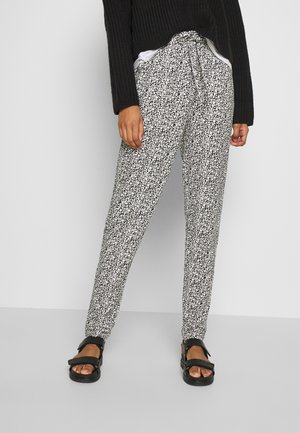 Persoon die zwart-witte broek met bloemenprint, een zwarte trui en zwarte sandalen draagt, staand tegen een effen lichte achtergrond.