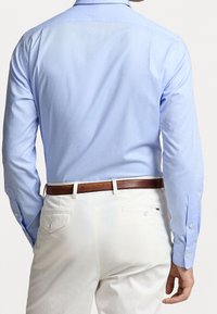 Camisa de manga larga azul claro con puños abotonados y cuello recto, combinada con pantalones blancos y un cinturón de cuero marrón.
