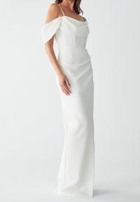Donna che indossa un elegante abito bianco off-shoulder con dettagli drappeggiati e sottili spalline, in piedi contro uno sfondo chiaro e uniforme.