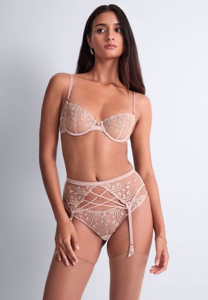 Femme aux longs cheveux noirs portant un ensemble de lingerie beige transparent brodé avec jarretelles et bas, sur un fond clair uni.