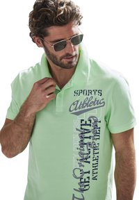 Hellgrünes Polo-Shirt mit kurzen Ärmeln, auf der Vorderseite mit großem, gedrucktem Text in Blau und Dunkelgrau versehen. Der Stoff wirkt glatt.