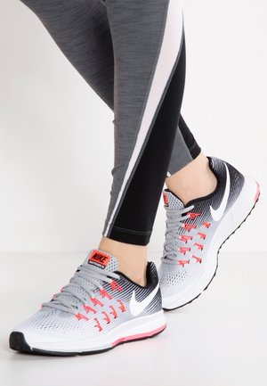 Nohy v šedých a černých běžeckých botách Nike s červenými detaily a šedými tkaničkami, v kombinaci se šedými a černými legíny s bílými pruhy.